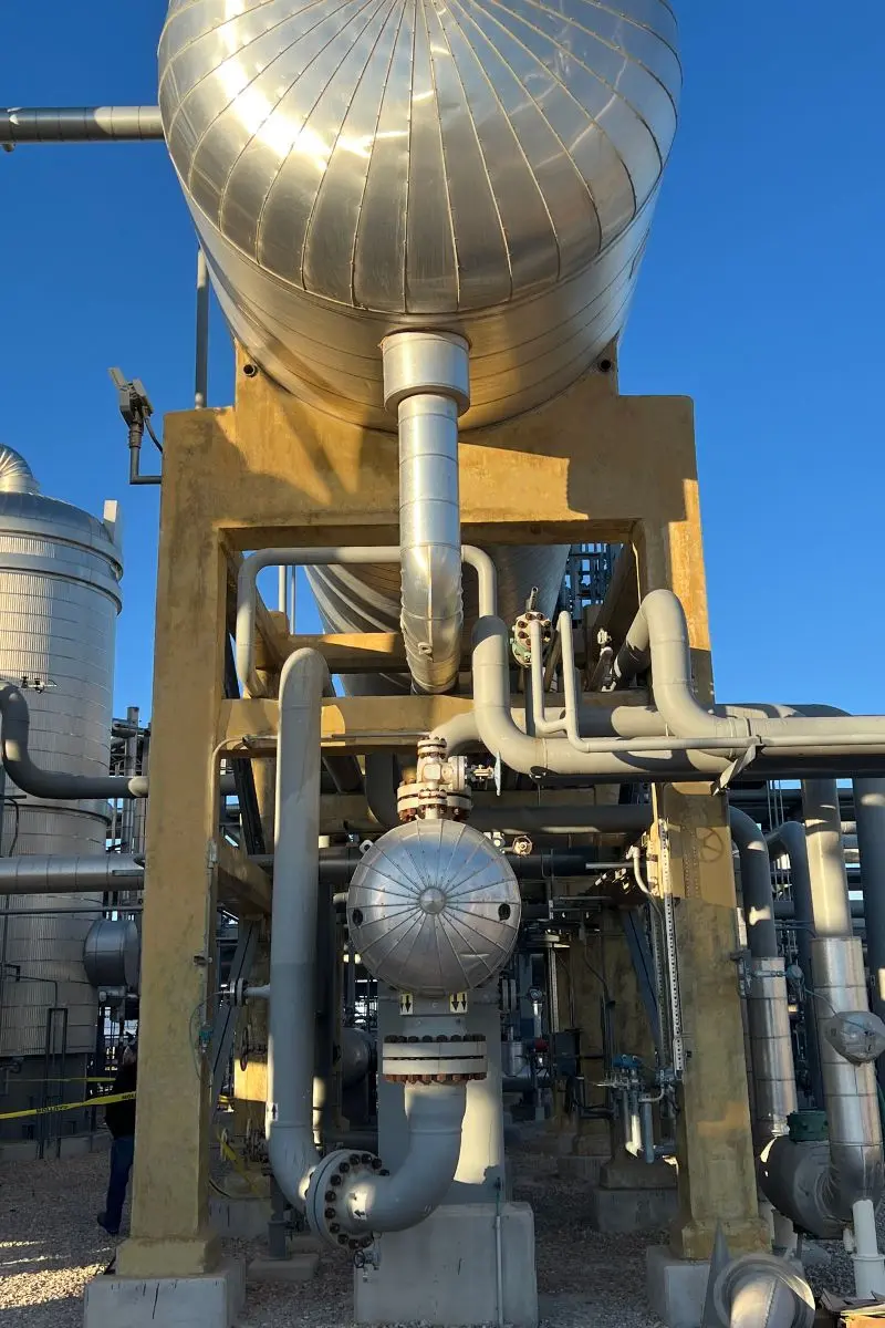 Large industrial storage tank with multiple pipes, valves, and metal structures under a clear blue sky at an industrial facility utilizing advanced Geopolymer Solutions.